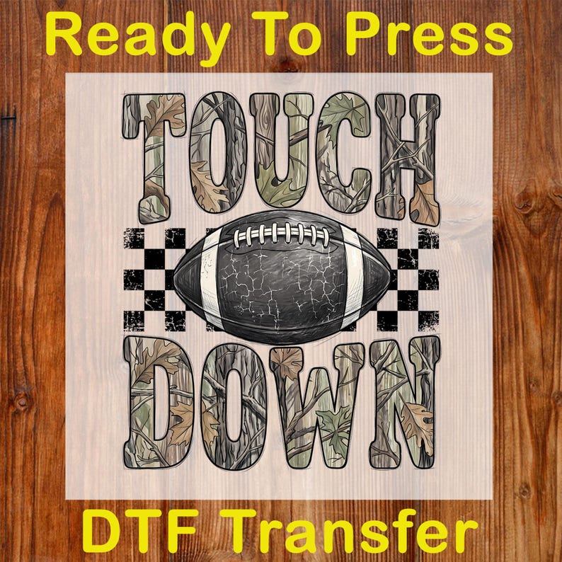 Pode incluir: Uma folha de transfer&ecirc;ncia DTF com o texto TOUCH DOWN num padr&atilde;o de folhas de camuflagem. Uma bola de futebol americano preta com atacadores brancos e textura rachada est&aacute; entre as palavras, sobre um padr&atilde;o de xadrez preto e branco.