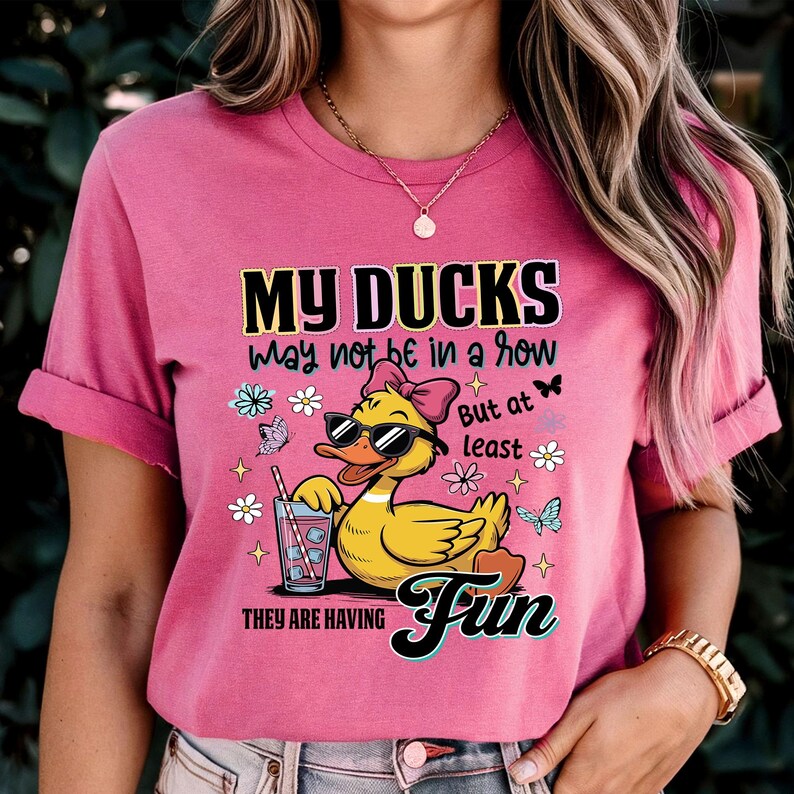 May include: Pink t-shirt with the text "MY DUCKS may not be in a row But at least THEY ARE HAVING Fun". The graphic features a yellow duck wearing sunglasses and a bow, holding a drink with a straw, surrounded by flowers and butterflies.