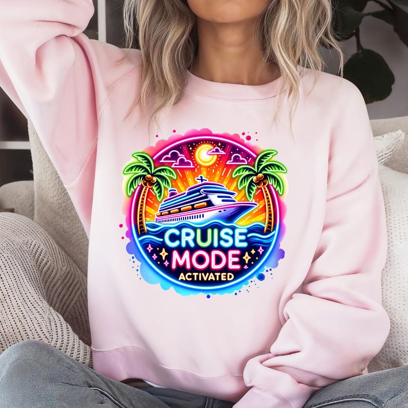 May include: A light pink sweatshirt featuring a colorful graphic with a cruise ship, palm trees, and the words "CRUISE MODE ACTIVATED." The design includes a sunset, moon, and stars.