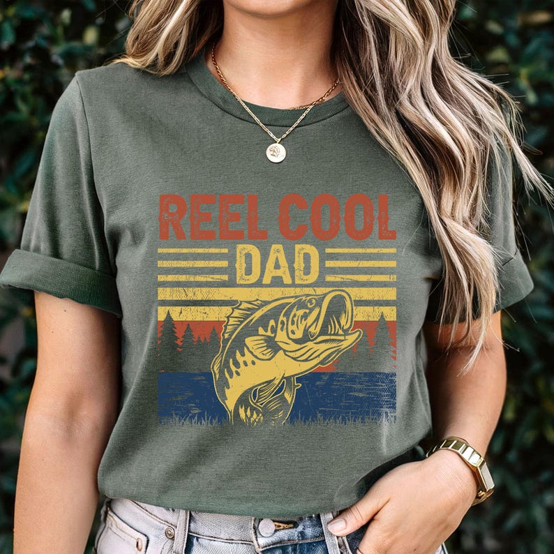 May include: Olive green t-shirt with the text "REEL COOL DAD" in red and yellow. Below the text is a graphic of a fish jumping out of the water, with a forest and water background. The shirt is a casual style.