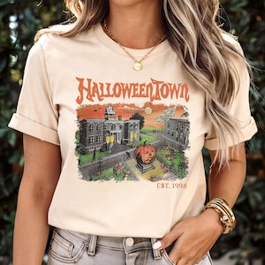 May include: Beige t-shirt featuring a Halloween Town graphic. The design includes a pumpkin, buildings, and the text "HALLOWEENTOWN EST. 1998" in orange. The shirt has short sleeves and a crew neck.