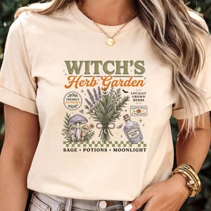 May include: A cream-colored t-shirt featuring a graphic print with the text "WITCH'S Herb Garden" and "SAGE &bull; POTIONS &bull; MOONLIGHT". The design includes illustrations of a lavender bouquet, mushrooms, a potion bottle, and small stars.