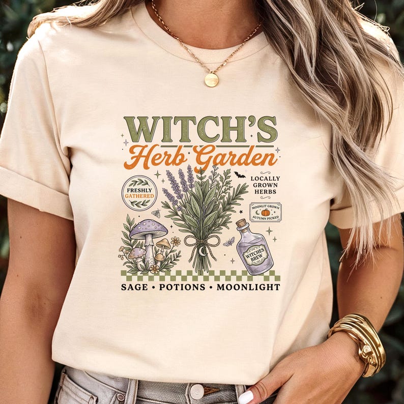 May include: A cream-colored t-shirt featuring a graphic print with the text "WITCH'S Herb Garden" and "SAGE &bull; POTIONS &bull; MOONLIGHT". The design includes illustrations of a lavender bouquet, mushrooms, a potion bottle, and small stars.