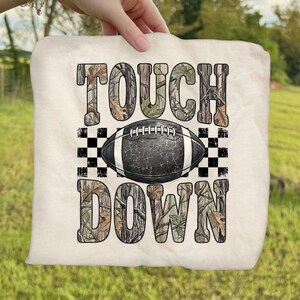 Pode incluir: Um item de tecido na cor creme com o texto TOUCH DOWN em um padr&atilde;o de camuflagem. Centralizada entre as palavras est&aacute; uma bola de futebol americano preta com cadar&ccedil;os brancos e textura rachada, ladeada por quadrados xadrez pretos e brancos.