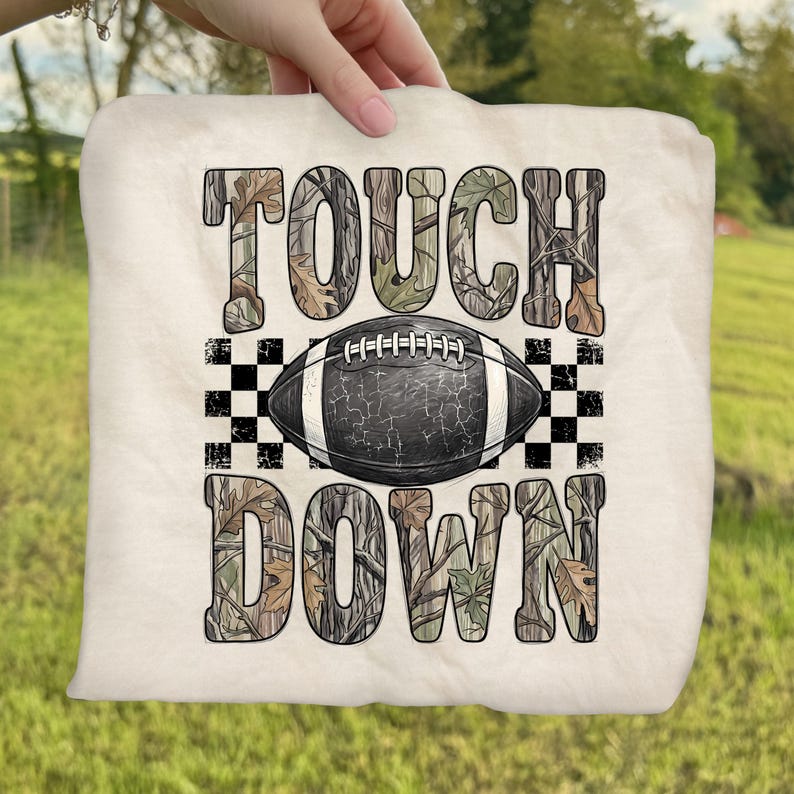 Pode incluir: Um item de tecido na cor creme com o texto TOUCH DOWN em um padr&atilde;o de camuflagem. Centralizada entre as palavras est&aacute; uma bola de futebol americano preta com cadar&ccedil;os brancos e textura rachada, ladeada por quadrados xadrez pretos e brancos.