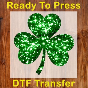 May include: A green glitter shamrock design, ready to press, with the text "Ready To Press" and "DTF Transfer" in yellow. The shamrock is a three-leaf clover shape, perfect for St. Patrick's Day crafts.