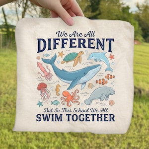 May include: Cream-colored tote bag featuring a colorful illustration of various sea creatures, including a whale, dolphin, and octopus. The text reads "We Are All DIFFERENT But In This School We All SWIM TOGETHER."