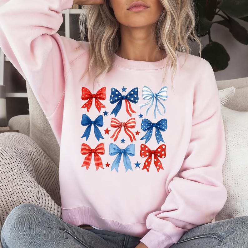 May include: A light pink sweatshirt featuring a pattern of nine bows in red, white, and blue. The bows are in various styles, with stars and stripes. The sweatshirt is a casual, comfortable style.