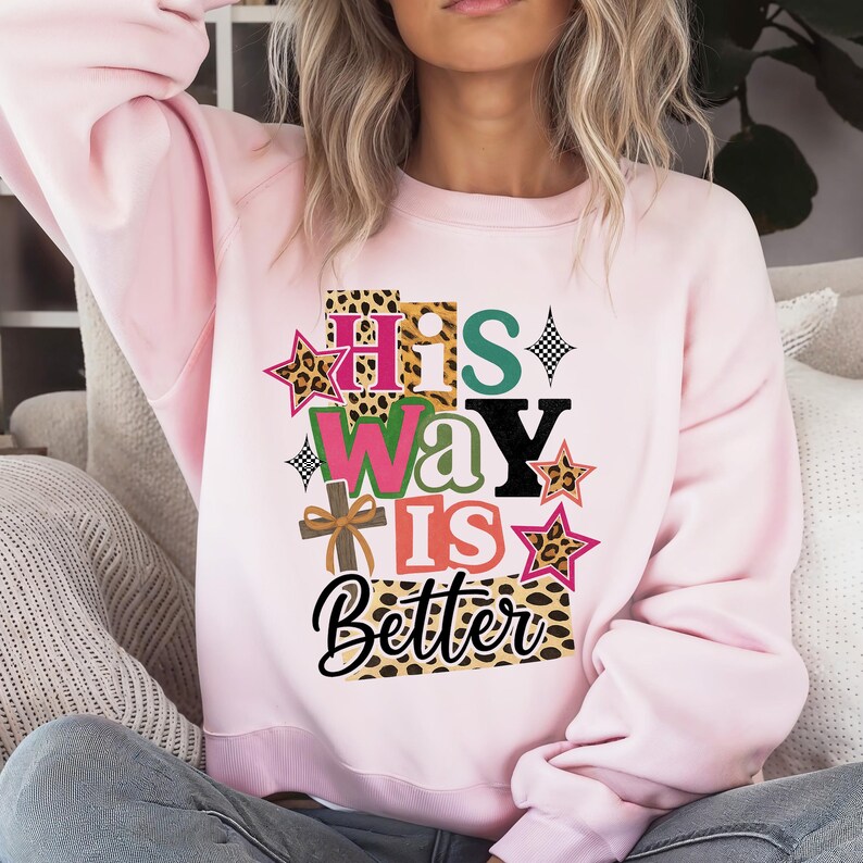 Op de afbeelding: Een lichtroze sweatshirt met de tekst "His Way Is Better" in een kleurrijk, gelaagd ontwerp. De tekst bevat luipaardprint accenten, sterren en een kruisafbeelding. De sweatshirt is een casual, comfortabele stijl.