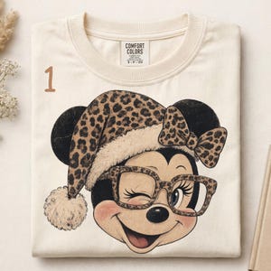 Puede incluir: Una camiseta color crema con el gráfico de un ratón de dibujos animados que lleva un gorro de Papá Noel con estampado de leopardo y gafas a juego. El ratón guiña un ojo. La camiseta tiene una etiqueta Comfort Colors y el número 1 en marrón.