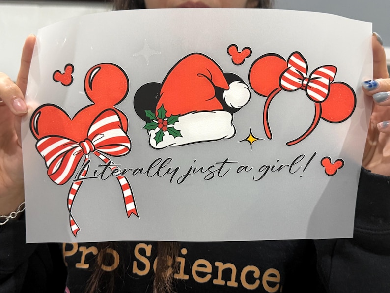 Puede incluir: Una l&aacute;mina transparente con dise&ntilde;os rojos y blancos con tem&aacute;tica de Mickey Mouse. Incluye un lazo, un gorro de Pap&aacute; Noel y orejas de Minnie Mouse. El texto "Literally just a girl!" est&aacute; escrito en cursiva. Tambi&eacute;n hay una estrella dorada e iconos de Mickey Mouse.