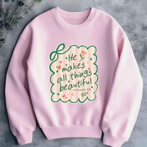 May include: A light pink sweatshirt with a crew neck. The front features a decorative design with the text "He makes all things beautiful" in a whimsical font, surrounded by hearts, stars, and floral elements. The text "ecclesiastes 3:16" is also included.
