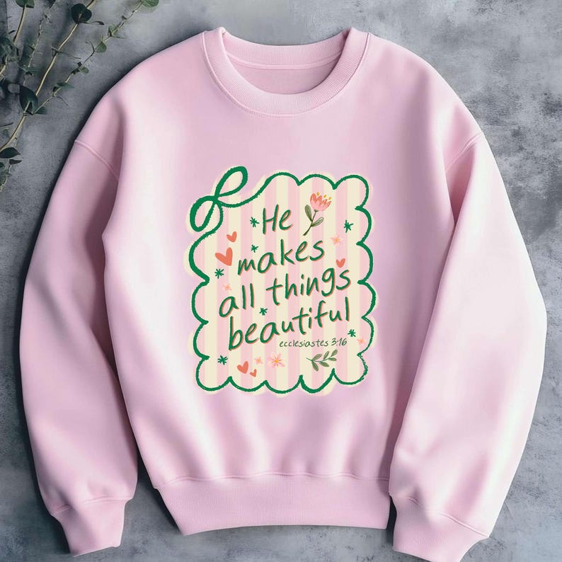 May include: A light pink sweatshirt with a crew neck. The front features a decorative design with the text "He makes all things beautiful" in a whimsical font, surrounded by hearts, stars, and floral elements. The text "ecclesiastes 3:16" is also included.