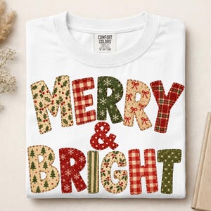 May include: A white crew neck t-shirt featuring the text MERRY & BRIGHT in large, multi-patterned letters. The letters display various holiday-themed prints including red and white gingham, green with snowflakes, holly berries, and pine trees.