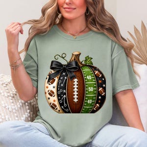 May include: A sage green t-shirt featuring a large graphic of a pumpkin designed with football elements. The pumpkin includes a black bow, a brown football texture, a green field with yard lines, and white play diagrams with small footballs.