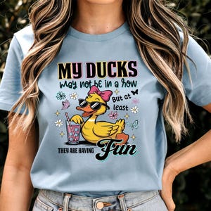 May include: Light blue t-shirt featuring a cartoon duck wearing sunglasses and a pink bow, holding a drink with a straw. The shirt has the text "MY DUCKS may not be in a row But at least THEY ARE HAVING Fun" in a colorful design.