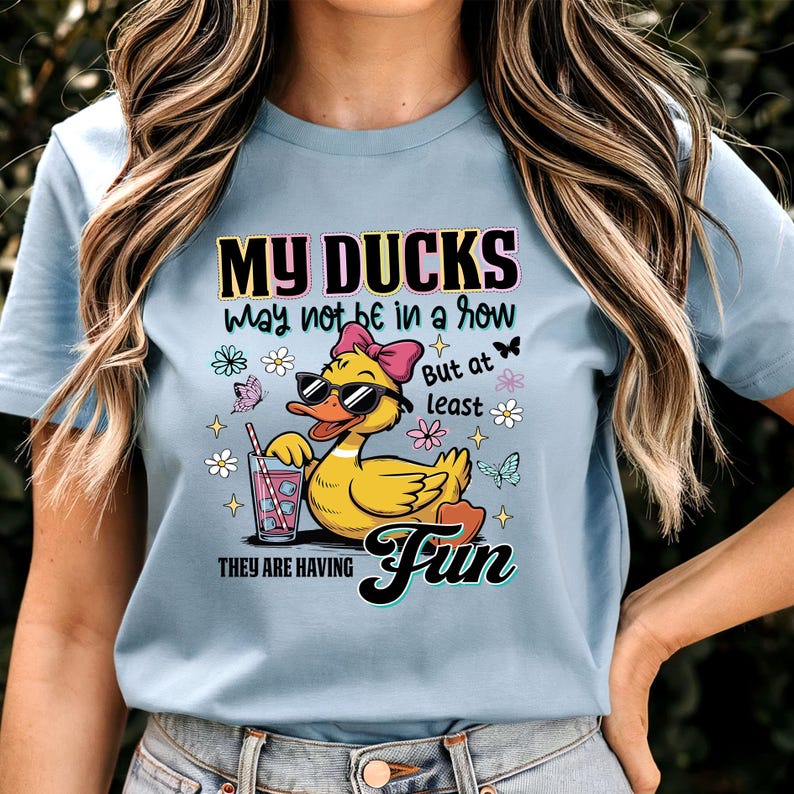 May include: Light blue t-shirt featuring a cartoon duck wearing sunglasses and a pink bow, holding a drink with a straw. The shirt has the text "MY DUCKS may not be in a row But at least THEY ARE HAVING Fun" in a colorful design.
