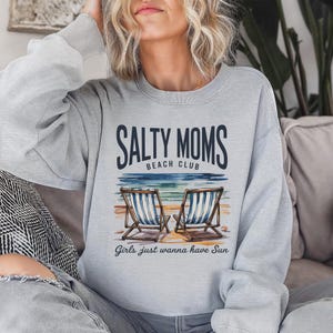 May include: A pale grey sweatshirt featuring the text "Salty Moms Beach Club" and an image of two beach chairs. The text "Girls just wanna have Sun" is below the image. The sweatshirt has a crew neck design.