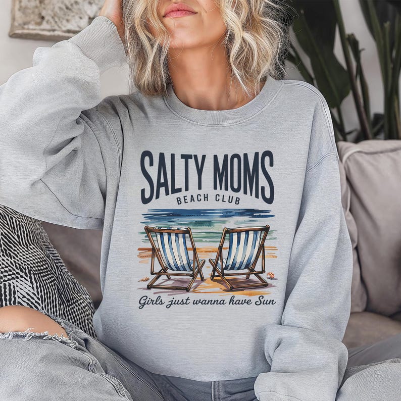May include: A pale grey sweatshirt featuring the text "Salty Moms Beach Club" and an image of two beach chairs. The text "Girls just wanna have Sun" is below the image. The sweatshirt has a crew neck design.