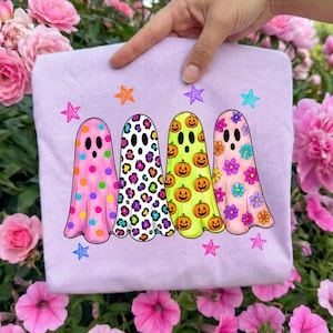 May include: A light purple t-shirt featuring a graphic of four ghosts standing in a row. The first ghost has pink polka dots, the second has a colorful leopard print, the third is lime green with orange pumpkins, and the fourth has floral patterns.