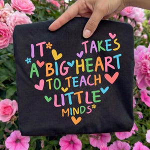 May include: Black t-shirt with the text "It takes a big heart to teach little minds" in colorful letters and heart shapes. The shirt is held up against a backdrop of pink flowers.