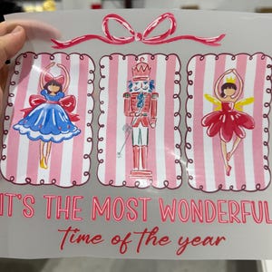 Op de afbeelding: Een transparante folie met drie feestelijke illustraties: een ballerina in een blauwe jurk, een notenkraker en een ballerina in een rode jurk. De tekst "IT'S THE MOST WONDERFUL Time of the year" staat in rode letters eronder. Bovenop zit een roze strik.