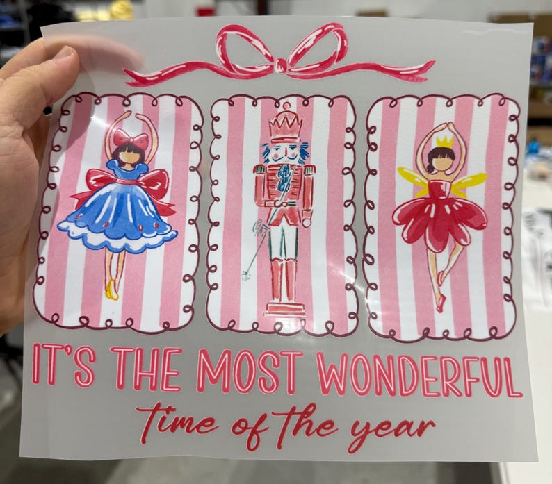 Op de afbeelding: Een transparante folie met drie feestelijke illustraties: een ballerina in een blauwe jurk, een notenkraker en een ballerina in een rode jurk. De tekst "IT'S THE MOST WONDERFUL Time of the year" staat in rode letters eronder. Bovenop zit een roze strik.