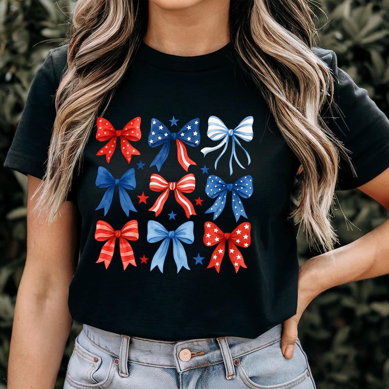 May include: Black t-shirt featuring a pattern of red, white, and blue bows. The bows have various designs, including stars and stripes. The shirt is a crew neck and is made of a soft material.