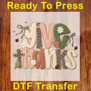 May include: A DTF transfer with the words "GIVE THANKS" in a decorative, stitched design. The letters feature a mix of colors, including cream, pink, green, and brown, with leopard print accents. The image includes floral and leaf embellishments.