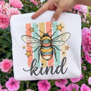 May include: White sweatshirt featuring a colorful bee illustration with the word "kind" in a script font. The bee is detailed with yellow, black, and blue stripes, surrounded by stars and vertical stripes in pastel colors.
