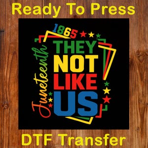 May include: A black DTF transfer with the words "THEY NOT LIKE US" in yellow, red, and blue. The word "Juneteenth" is in green script. The number "1865" is in green and red with stars. The text "Ready To Press" and "DTF Transfer" are in yellow.
