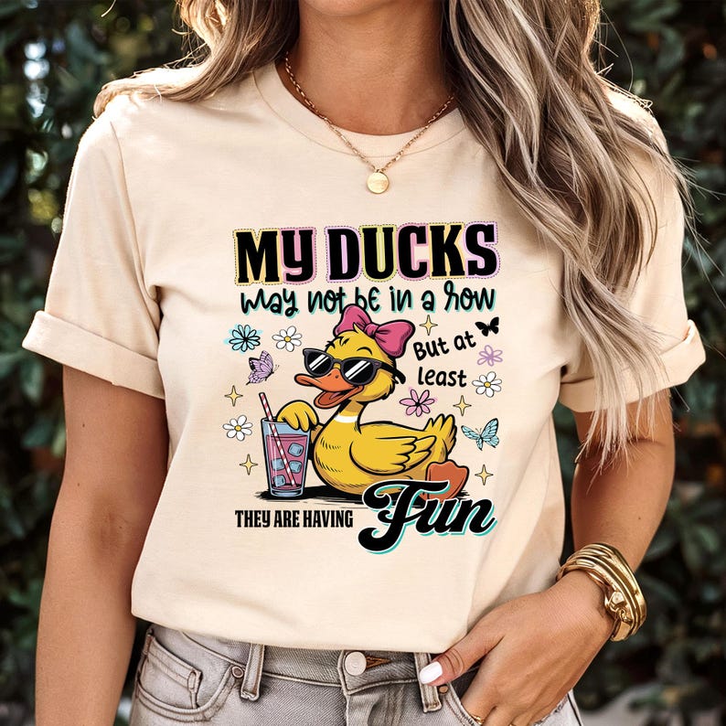 May include: Beige t-shirt with the text "MY DUCKS may not be in a row But at least THEY ARE HAVING Fun." The graphic features a cartoon duck wearing sunglasses and a pink bow, holding a drink with a straw. Butterflies and flowers surround the duck.