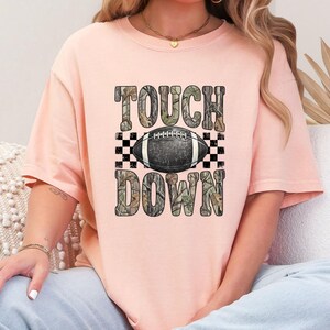 Pode incluir: Uma t-shirt de manga curta cor de p&ecirc;ssego com uma impress&atilde;o gr&aacute;fica com o texto TOUCH DOWN num padr&atilde;o de camuflagem. Entre as palavras encontra-se uma bola de futebol americano preta com riscas brancas sobre um fundo axadrezado.