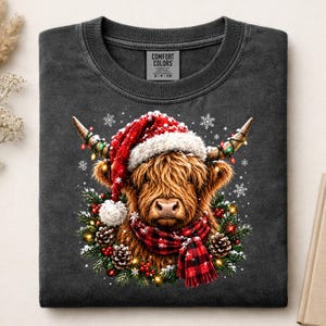 May include: A charcoal gray crewneck t-shirt featuring a graphic of a Highland cow wearing a red and white Santa hat and a red and black plaid scarf. The cow is surrounded by a wreath of pine branches, pinecones, red berries, and string lights.