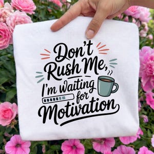 May include: A white sweatshirt held by a hand, featuring the text "Don't Rush Me, I'm waiting for Motivation" in black, teal, and orange. A coffee cup graphic is also included. The background is blurred with pink flowers.