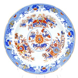 May include: A round ceramic plate featuring a white base with a central floral motif in orange, red, and blue. The border is decorated with intricate blue and white patterns, interspersed with orange and blue floral and butterfly accents.