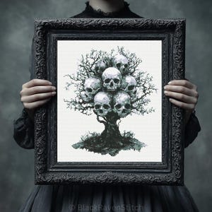 Può includere: Un'opera d'arte a punto croce incorniciata raffigurante un albero con teschi al posto delle foglie. L'opera è in tonalità di grigio e bianco, su uno sfondo neutro. La cornice è nera e ornata.