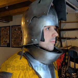 K&ouml;nnte beinhalten: Ein polierter mittelalterlicher Helm aus Stahl mit Visier und Nackenschutz. Der Helm wird &uuml;ber einem gelben Kleidungsst&uuml;ck getragen. Das Bild zeigt ein Seitenprofil des Helms und der Person, die ihn tr&auml;gt.