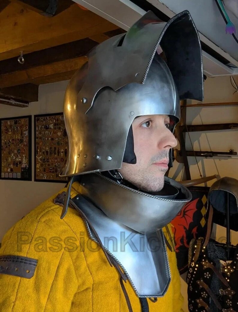 K&ouml;nnte beinhalten: Ein polierter mittelalterlicher Helm aus Stahl mit Visier und Nackenschutz. Der Helm wird &uuml;ber einem gelben Kleidungsst&uuml;ck getragen. Das Bild zeigt ein Seitenprofil des Helms und der Person, die ihn tr&auml;gt.