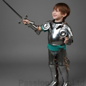 May include: A child in shiny silver armor, complete with a helmet, chest plate, arm guards, and leg armor, holds a silver sword. The armor is detailed with rivets and plates. The child is smiling and wearing a teal shirt.