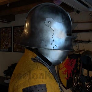 K&ouml;nnte beinhalten: Ein polierter Stahlhelm mit Visier, Teil einer R&uuml;stung. Der Helm hat eine abgerundete Oberseite und ein gebogenes Visier mit Nieten. Der Helm wird &uuml;ber einem gelben Kleidungsst&uuml;ck mit einem dunkelgrauen Aufn&auml;her am &Auml;rmel getragen. Im Hintergrund befinden sich weitere mittelalterliche Gegenst&auml;nde.