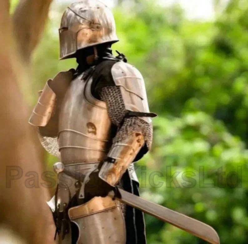 Puede incluir: Una armadura medieval, que incluye un casco, una placa para el pecho y protectores de brazos. La armadura es de metal y presenta cota de malla en los brazos. Una espada se sostiene en la mano derecha. El fondo est&aacute; borroso con follaje verde.