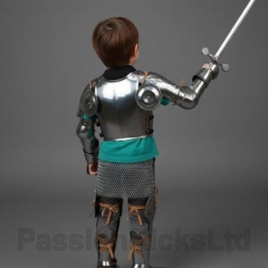 May include: A child wearing a full suit of silver-colored knight armor, including a breastplate, arm guards, chainmail, and leg armor. The child is holding a silver sword with a detailed hilt. The outfit is a costume, likely for play or a themed event.