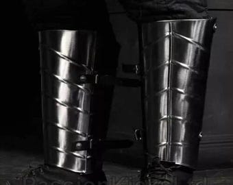 May include: Pair of polished metal greaves, part of leg armour, with a riveted design and leather straps. The armour is cylindrical and covers the lower leg, reflecting light. The image is in black and white.