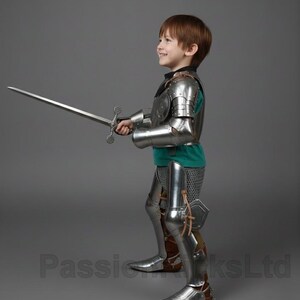 May include: A child in full knight's armor, including a breastplate, arm and leg guards, and a helmet. The child is holding a long sword. The armor is silver-colored, and the child is wearing a green shirt underneath.