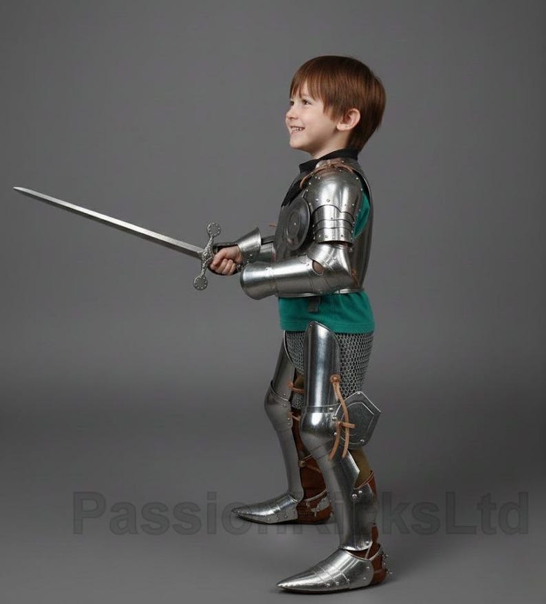May include: A child in full knight's armor, including a breastplate, arm and leg guards, and a helmet. The child is holding a long sword. The armor is silver-colored, and the child is wearing a green shirt underneath.