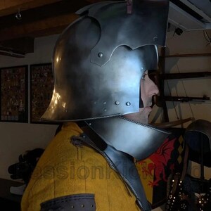 K&ouml;nnte beinhalten: Ein polierter mittelalterlicher Stahlhelm mit Visier und Nackenschutz. Der Helm hat Nieten und eine dekorative Oberseite. Die Person tr&auml;gt ein gelbes, gepolstertes Kleidungsst&uuml;ck. Der Hintergrund umfasst einen Schild und andere historische Gegenst&auml;nde.