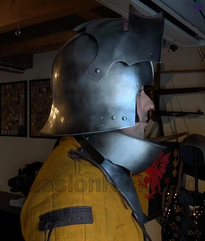 K&ouml;nnte beinhalten: Ein polierter mittelalterlicher Stahlhelm mit Visier und Nackenschutz. Der Helm hat Nieten und eine dekorative Oberseite. Die Person tr&auml;gt ein gelbes, gepolstertes Kleidungsst&uuml;ck. Der Hintergrund umfasst einen Schild und andere historische Gegenst&auml;nde.
