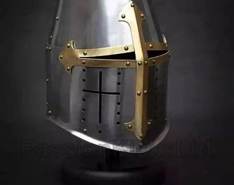 May include: A silver-colored medieval crusader helmet with a gold-colored cross design on the front. The helmet has a rounded top and is riveted. The helmet is displayed on a black stand.