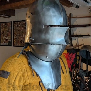 K&ouml;nnte beinhalten: Ein polierter mittelalterlicher Stahlhelm mit Visier und Nackenschutz, getragen &uuml;ber einem gelben, gepolsterten Kleidungsst&uuml;ck. Der Helm hat eine glatte, reflektierende Oberfl&auml;che. Das Kleidungsst&uuml;ck ist leuchtend gelb. Bild eines historischen oder Cosplay-Artikels.